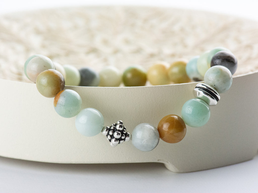 Black Gold Amazonite Genuine Gemstone Stretch Bracelet displayed on a decorative bowl. Amazonite beads are multicolor earth-tones. Center bead is a silver Bali style bead. Bracelet hangs slightly over side edge of decorative bowl.
