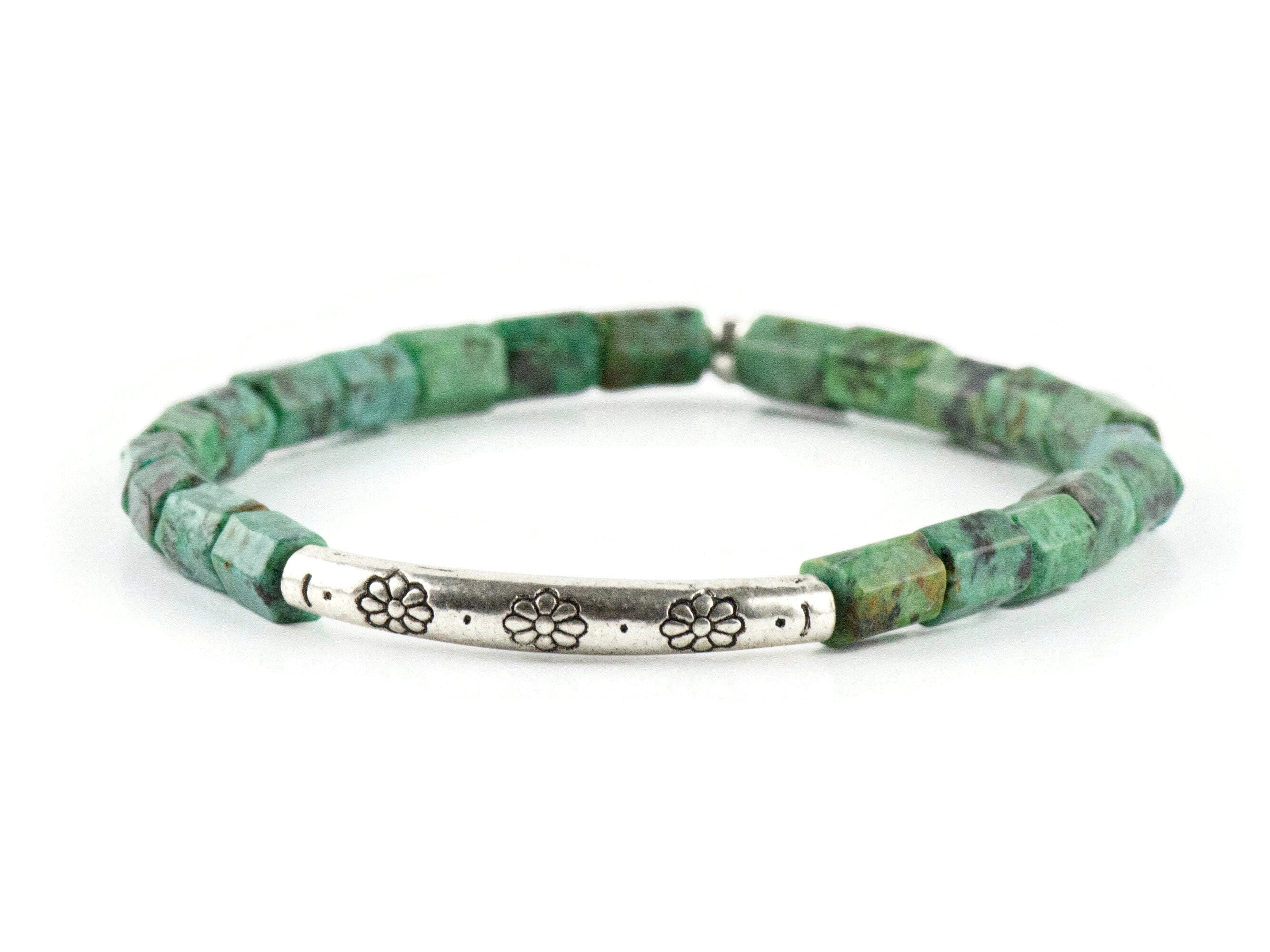 African Turquoise Stretch Bracelet on white background. Beads are bluish green with black and brown speckle. Silver pewter tube focal bead is adorned with floral etchings.